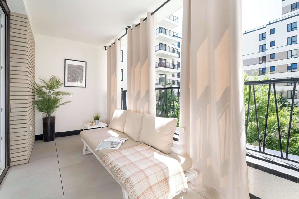 A long, narrow enclosed balcony offers a cozy relaxation spot with a padded bench and soft cushions. Glass windows overlook the neighborhood, flooding the space with natural daylight.