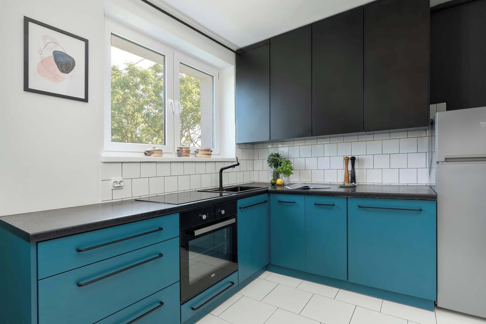 A modern L-shaped kitchen combines turquoise lower cabinets with white upper cupboards and a tiled backsplash. Large windows brighten the workspace throughout the day.