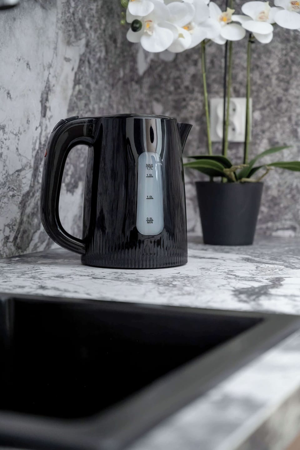 A detailed shot focuses on a sleek black electric kettle sitting on the marble countertop next to a delicate white orchid. The minimalist styling emphasizes functionality and elegance.