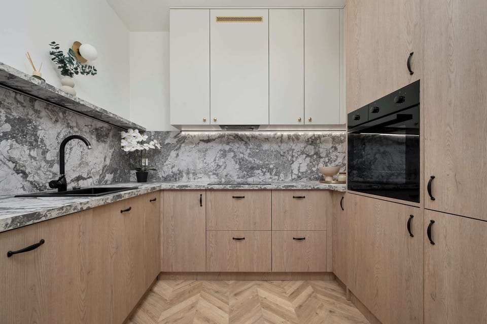 A full view of the modern kitchen highlights upper white cabinets contrasting gracefully with the lower wooden drawers. A built-in black oven is seamlessly integrated into the cabinetry next to the marble-patterned walls.