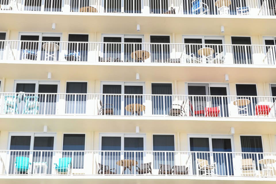 A beachfront property building featuring multiple floors with private balconies, white railings, and sliding glass doors leading to outdoor seating areas.