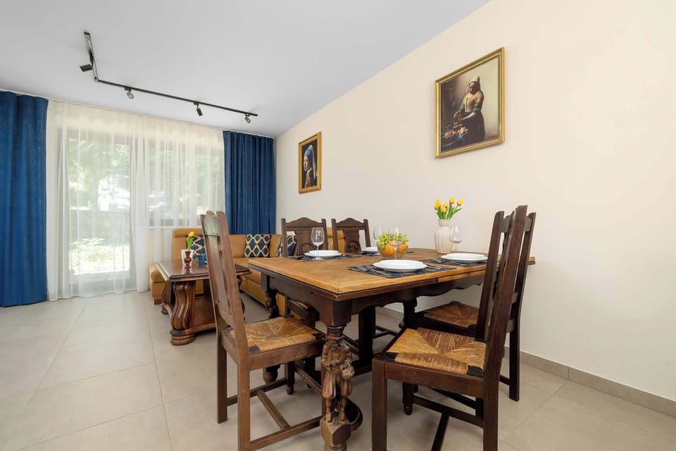 A massive rustic wooden dining table stands prominently in a spacious room, surrounded by six high-backed carved chairs. Large windows with blue drapes brighten the neutral walls.