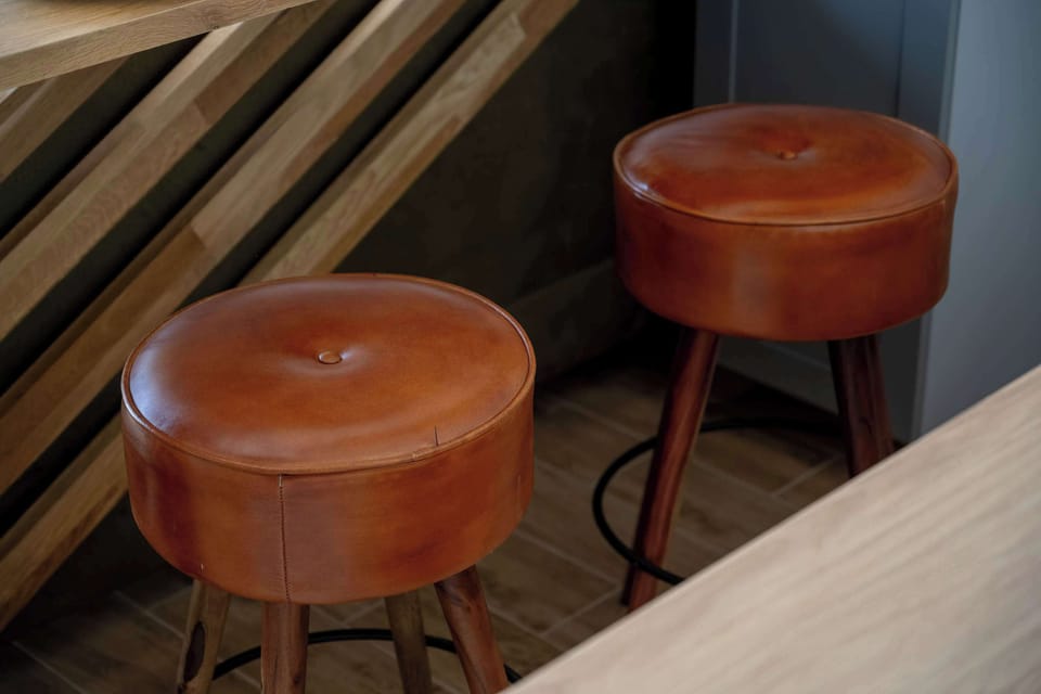 A close-up shot captures two modern bar stools with round, cognac-colored leather seats and dark metal legs. They are positioned neatly under the wooden countertop next to a slatted partition.
