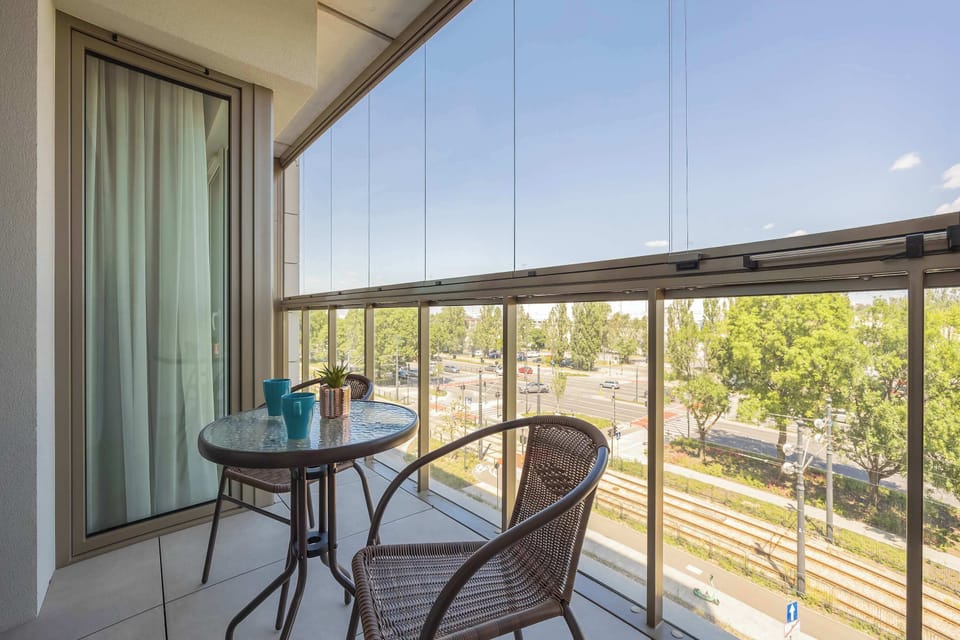 An enclosed glass loggia offers an elevated view of a green streetscape with tram lines below. A compact round table and a wicker armchair create a peaceful nook for outdoor relaxation.