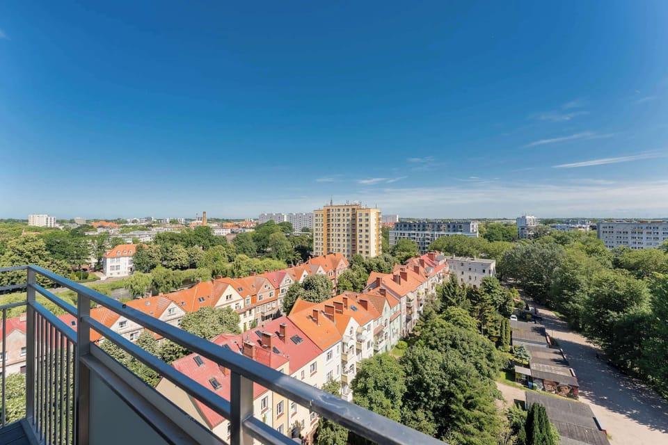 A panoramic perspective from the high-rise balcony showcases a vast expanse of green treetops extending toward the horizon. The bright blue sky creates a beautiful backdrop for the residential area below.