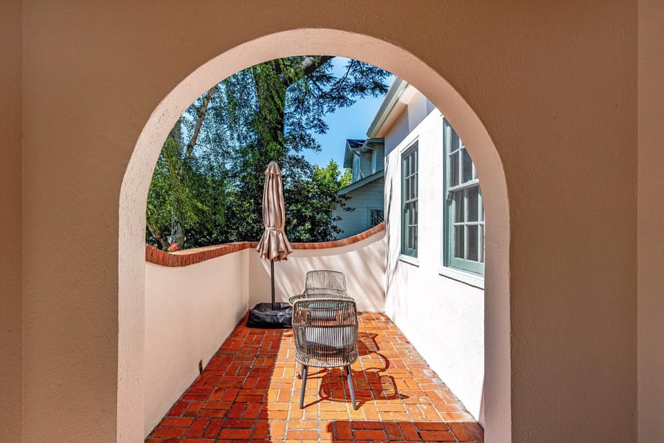 Your private courtyard invites peaceful mornings with warm terracotta tiles underfoot, stylish seating beneath a shaded umbrella, and lush greenery framing the sky above.