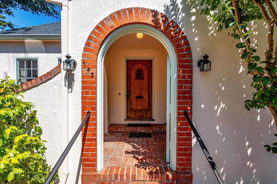Charming brick-arched entryway welcomes guests to this bright, sun-dappled property featuring a classic wooden door and inviting stucco exterior framed by lush greenery.