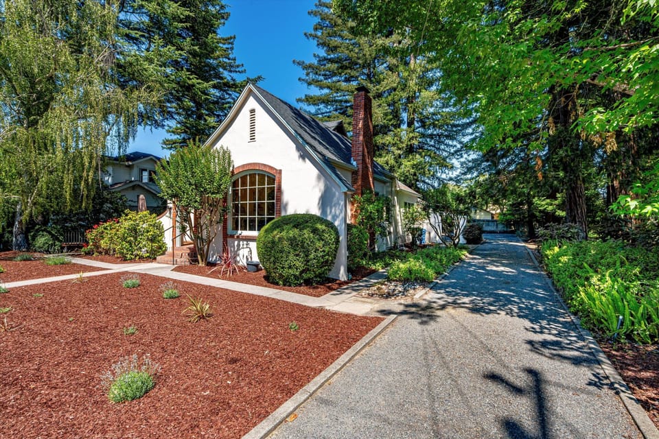 Charming white cottage with brick chimney and arched window, framed by towering evergreens and manicured landscaping along a private driveway.