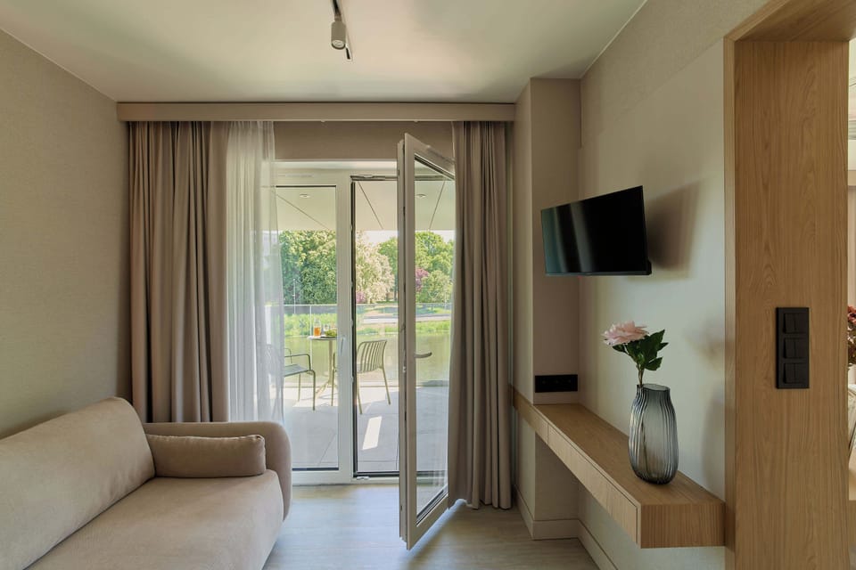 Salon został urządzony w nowoczesnym stylu, łączącym naturalne drewno, jasne beże i oliwkową zieleń. Znajduje się w nim wygodna, rozkładana sofa. Z pomieszczenia można wyjść bezpośrednio na balkon.