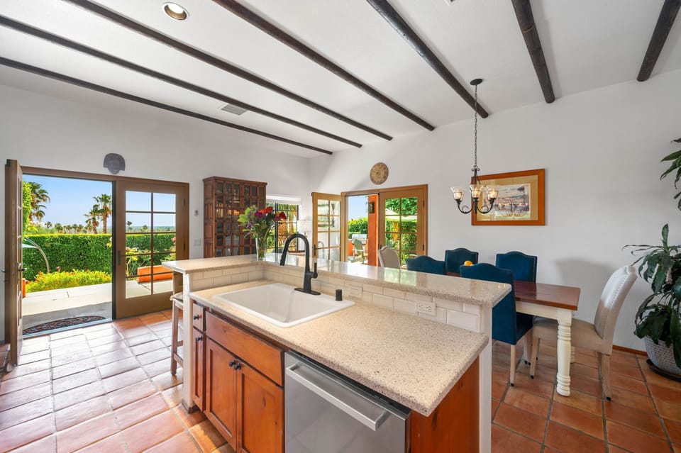 Your spacious kitchen opens to lush garden views, featuring a large island with modern sink, dining nook with blue cushioned seating, exposed beam ceilings, and terracotta tile floors creating a warm, inviting culinary retreat.