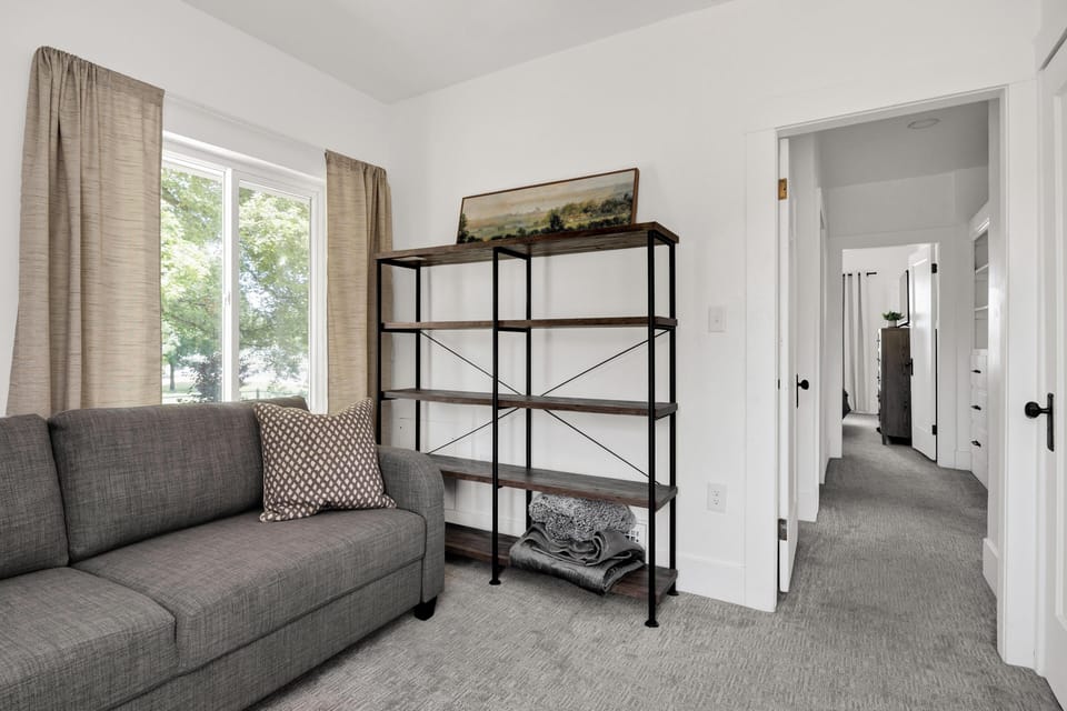 Gray sofa with patterned pillow sits beside black metal and wood shelving unit near beige curtains and window.