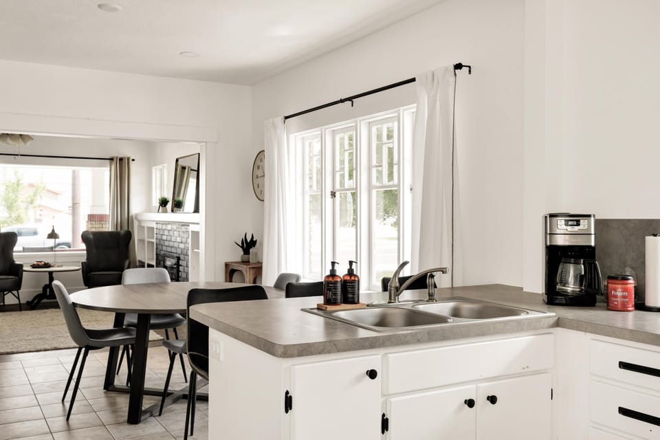 Concrete-look kitchen countertops frame a double stainless steel sink beside a black coffee maker and window with white curtains.