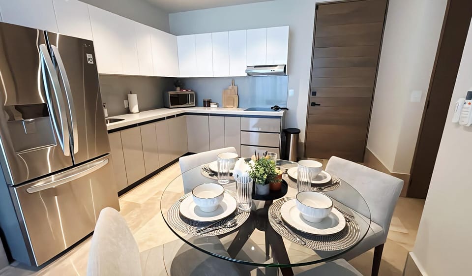 Glass-top dining table set for four with white cushioned chairs beside a sleek kitchen with stainless steel fridge.