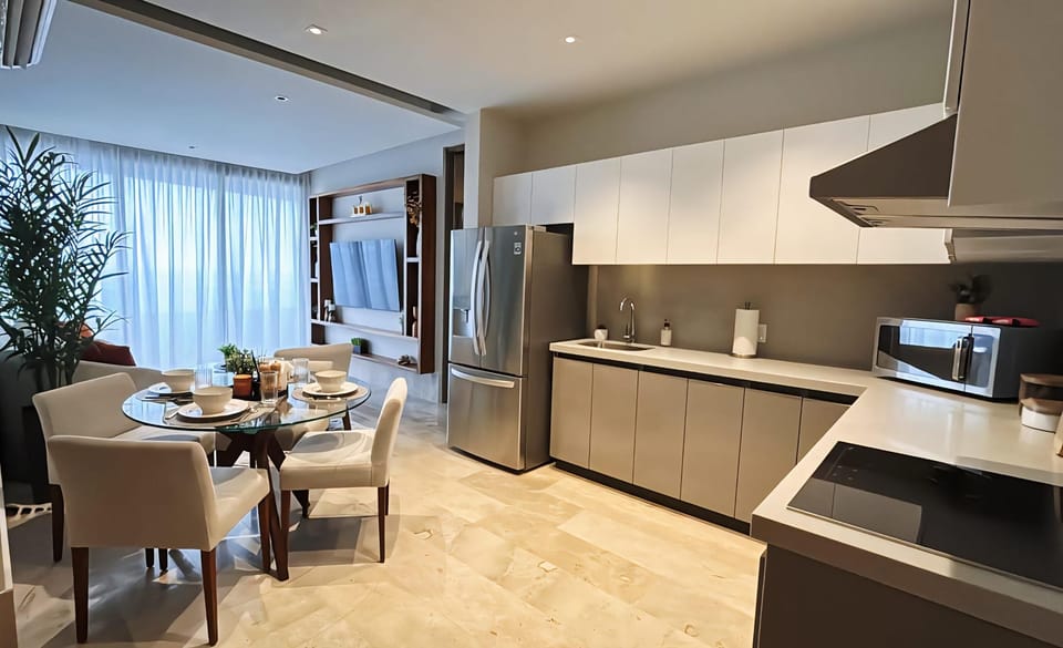 Glass-top dining table with white cushioned chairs sits adjacent to sleek kitchen cabinetry and stainless steel fridge.