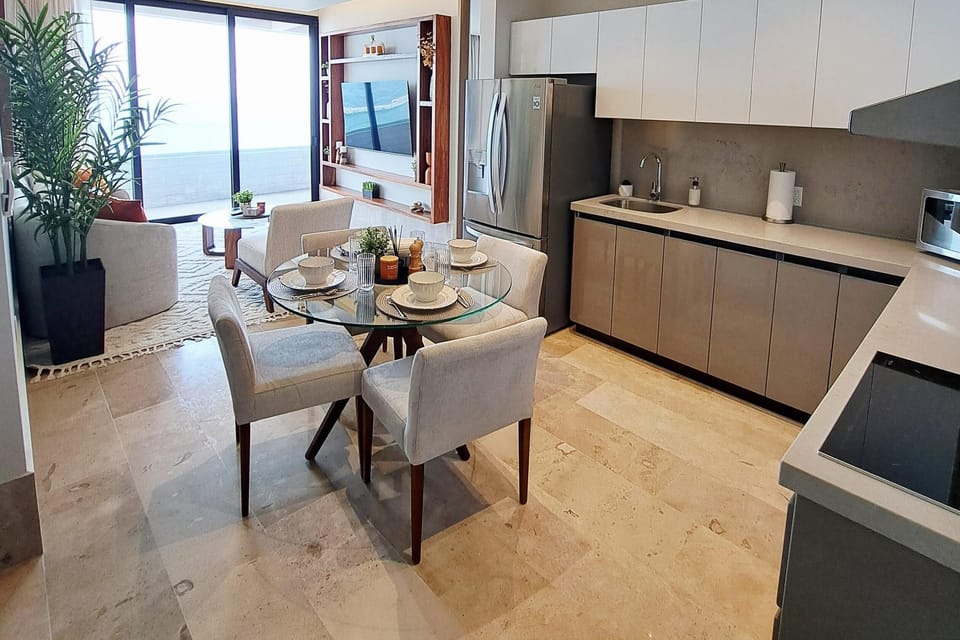 Glass dining table with four beige upholstered chairs sits on beige stone tile between kitchen and living area.