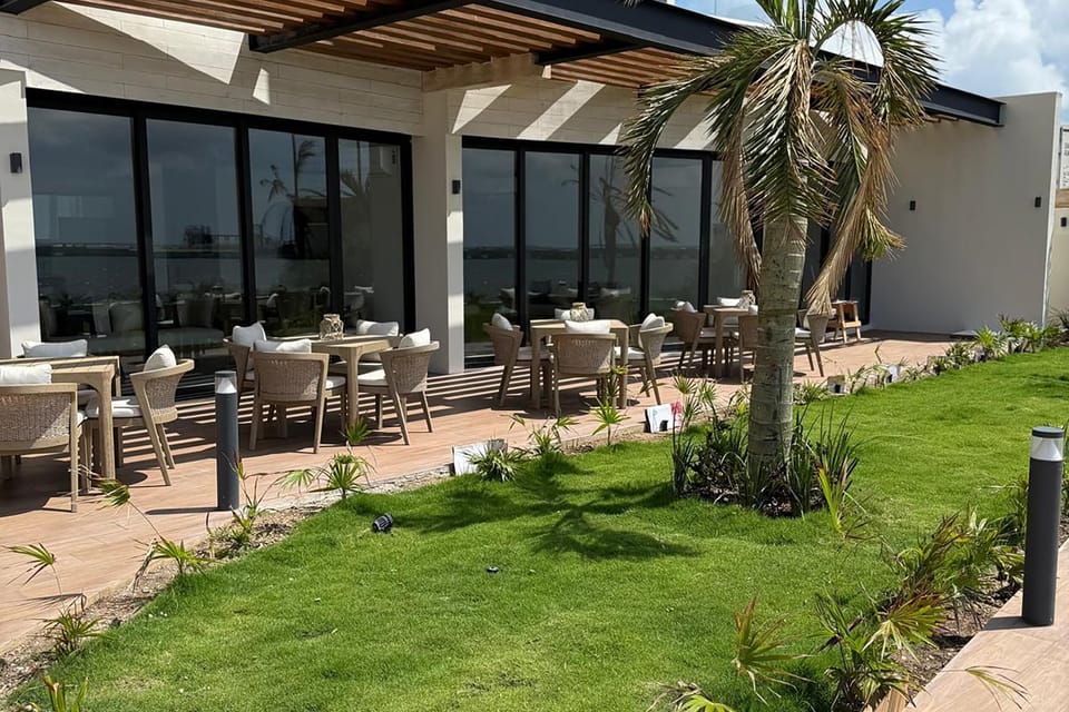 Outdoor patio with light wood tables, wicker chairs, white cushions, and a manicured lawn bordered by small plants.