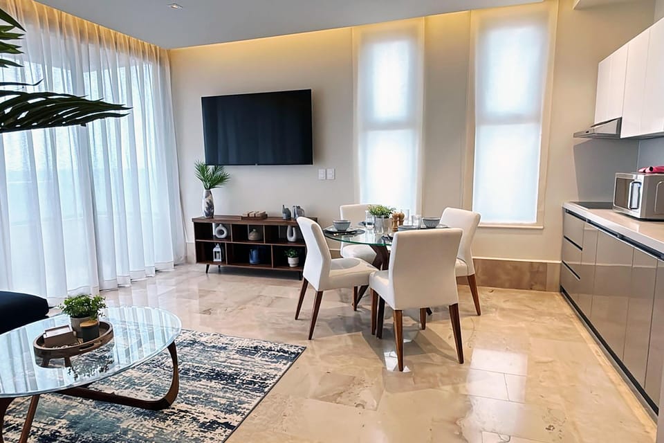 Open-concept living and dining area with marble floors, sheer curtains, and a wall-mounted flat-screen TV.