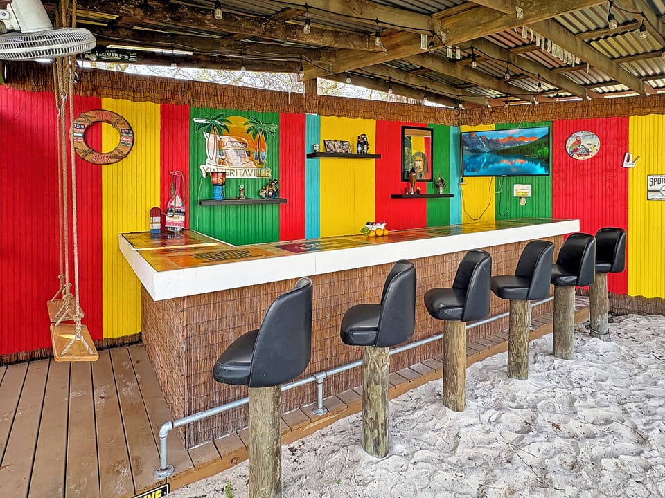 Colorful beachside tiki bar with sand-floor seating and TV lounge vibes