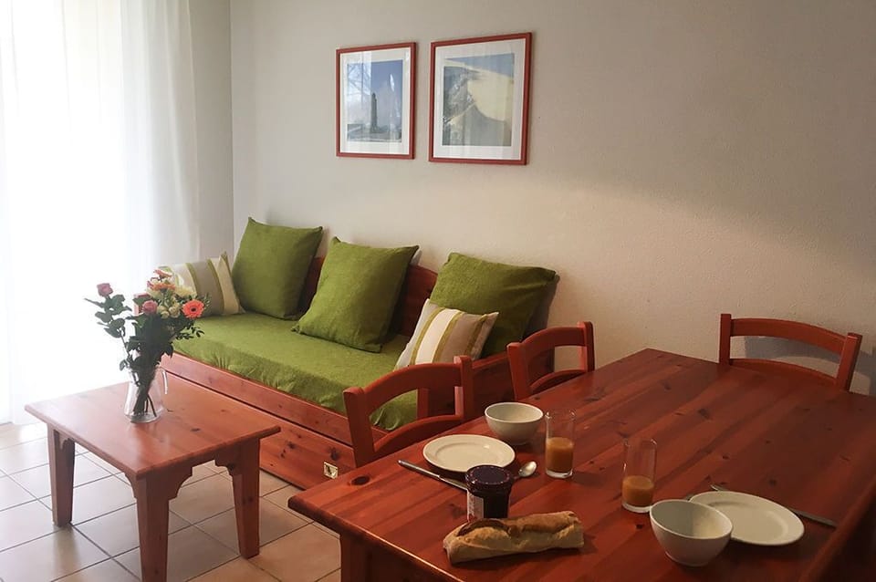 Dining space with wooden table and chairs, daybed with green cushions, and light-filled sheer curtains.