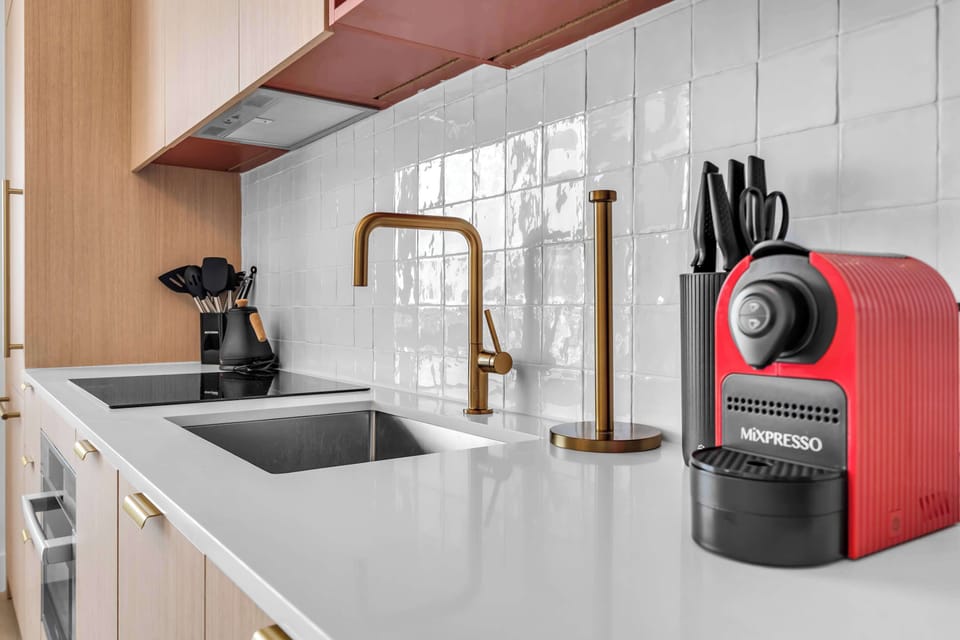 Close-up of a modern kitchen counter featuring a sleek electric cooktop, stainless steel sink, and elegant gold fixtures. Equipped with a premium red espresso machine and knife block, making your morning routine seamless and stylish.
