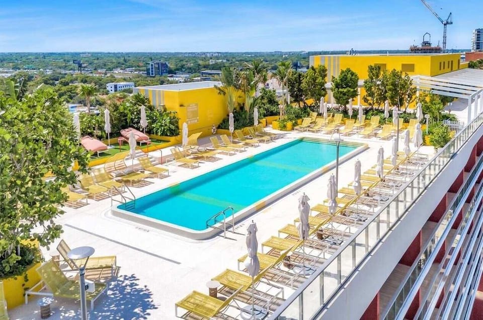 An aerial view of the spectacular resort-style rooftop swimming pool. Surrounded by rows of yellow lounge chairs, umbrellas, and lush greenery, this expansive sun deck offers the ultimate elevated oasis during your stay.