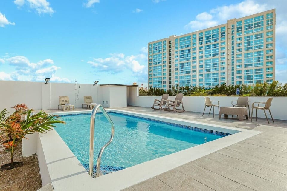 A rectangular turquoise swimming pool sits on a modern, light-gray tiled rooftop terrace. The deck is arranged with neutral loungers, rocking chairs, and patio seating under a bright, partly cloudy sky.