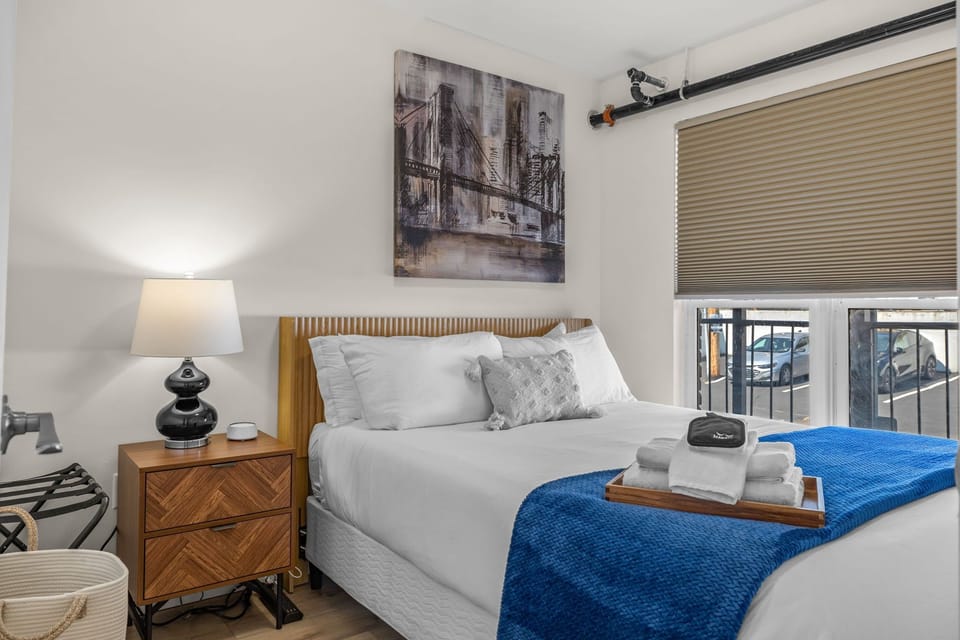 Bedroom with white linens, a vivid blue throw, wall art, bedside lamp and a window.