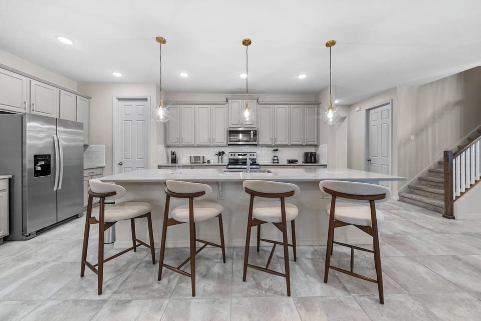 Welcome to a beautiful kitchen with sleek gray cabinetry, a spacious island with stylish bar seating, and modern pendant lights that accentuate the inviting marble-like flooring, creating a warm and sophisticated atmosphere for.