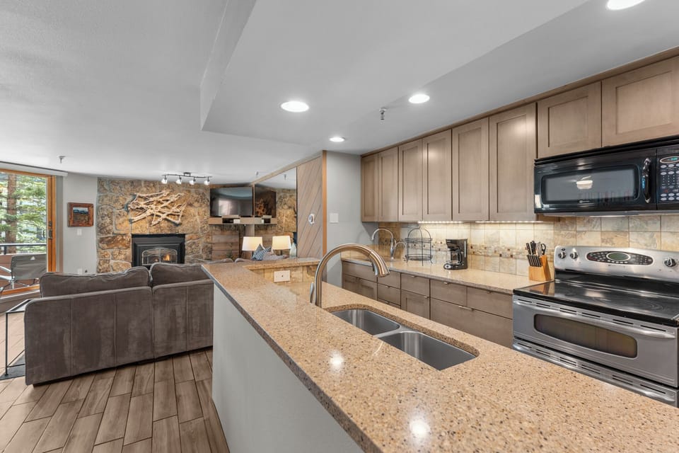 Your fully equipped kitchen opens to a cozy living room with a stone fireplace, perfect for preparing meals and relaxing together after mountain adventures.