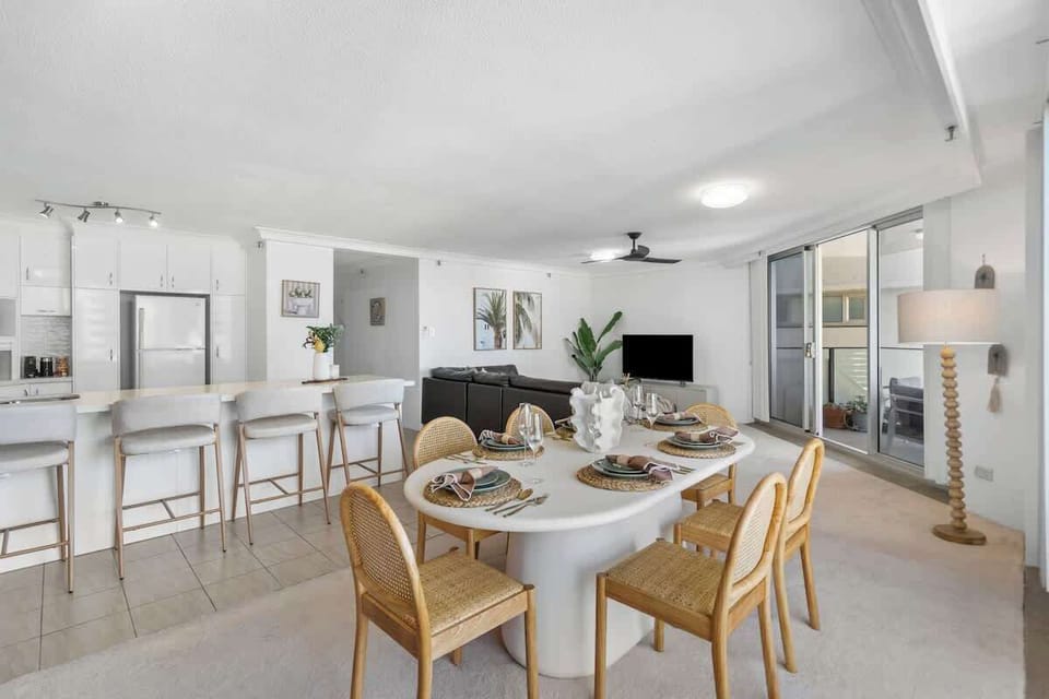 Experience open-concept coastal luxury in this spacious living and dining area, featuring an oval dining set, an expansive kitchen breakfast bar, and sliding glass doors that open to a breezy private balcony.