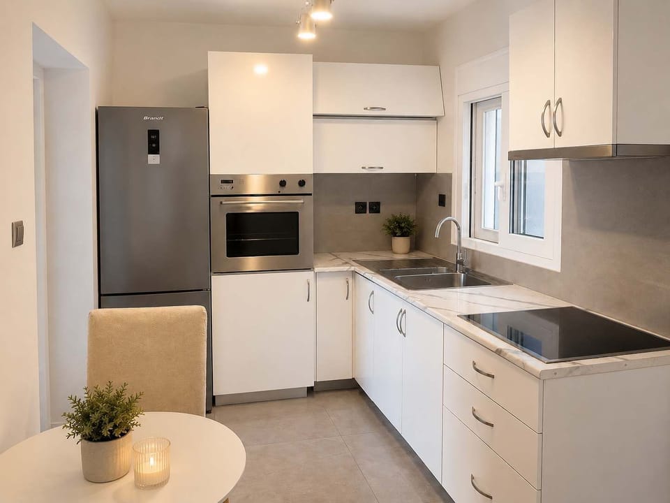 A compact modern kitchen with white cabinets, sleek appliances, marble-style counters, and a small dining corner. The clean lines and bright lighting make it feel fresh, practical, and inviting