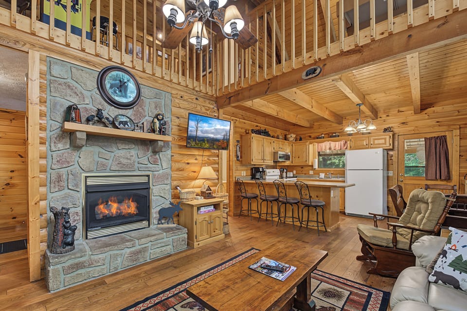 Unwind in your spacious cabin with soaring ceilings and a cozy stone fireplace glowing at the heart of the open living space, seamlessly blending rustic charm with modern comfort.