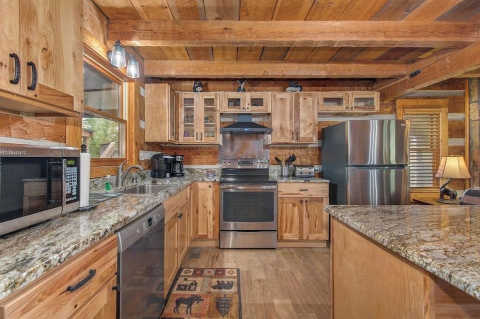 Your full cabin kitchen boasts granite counters, stainless appliances, and rustic wood beams overhead—a perfect blend of modern convenience and mountain charm for preparing meals together.