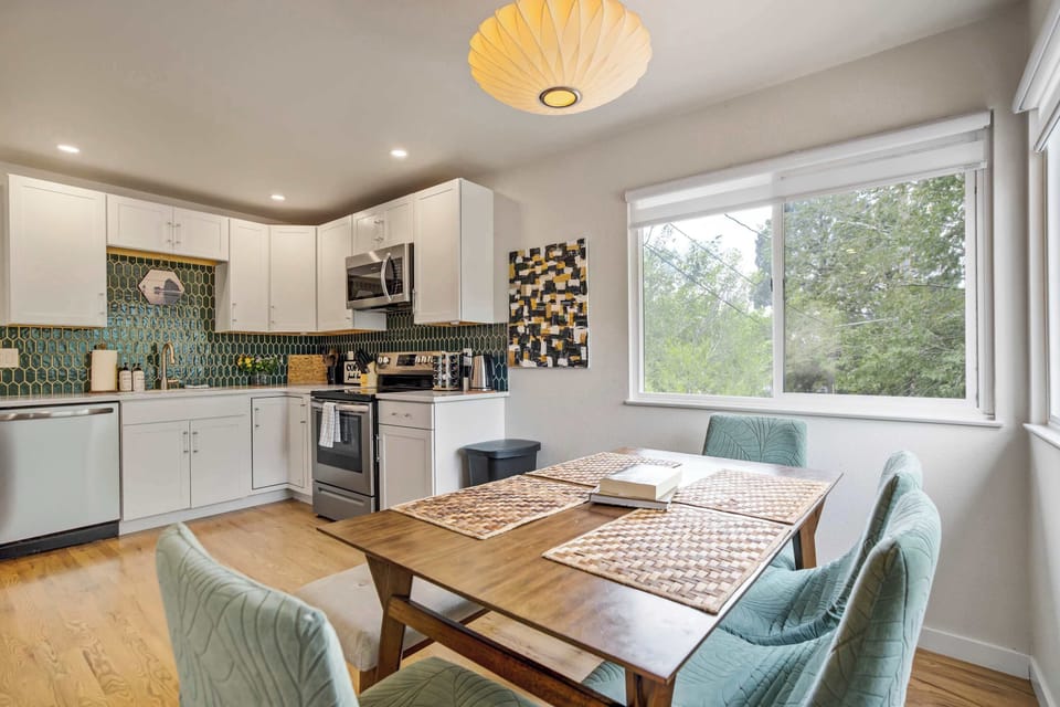 From the kitchen, the same corner window pulls tree lined views into view past the dining table, the mustard pendant glowing overhead while a mosaic tile accent adds a playful detail near the range.