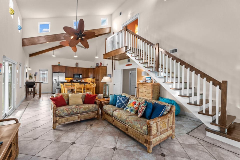 Open-concept living and dining room with vaulted ceilings, exposed beams, ceiling fan, tiled floors, and comfortable group seating.