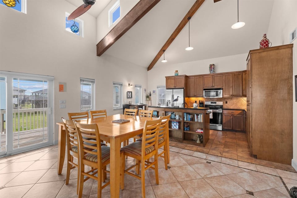 Dining area beside the kitchen with a large wood table, vaulted ceiling, bar seating, tiled flooring, and open coastal layout.