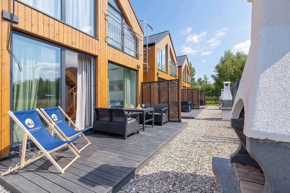 An outdoor terrace view of modern wooden cottages with blue deck chairs set out on the paved ground. The open doors hint at a seamless transition between indoor and outdoor living.