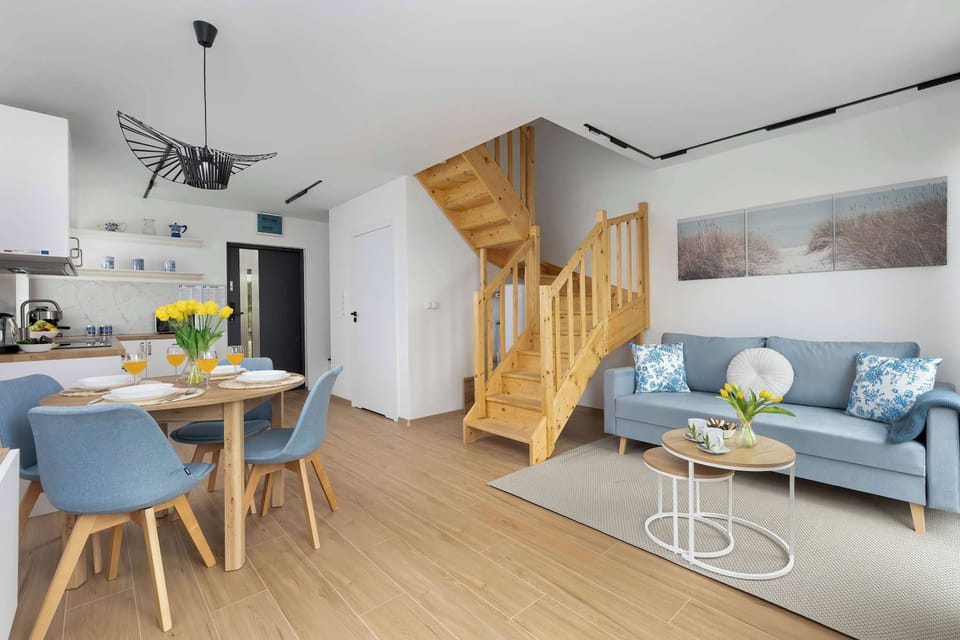 A bright living and dining combination with a blue sofa on the right and a dining table on the left. The wooden staircase serves as a beautiful architectural focal point in the room.