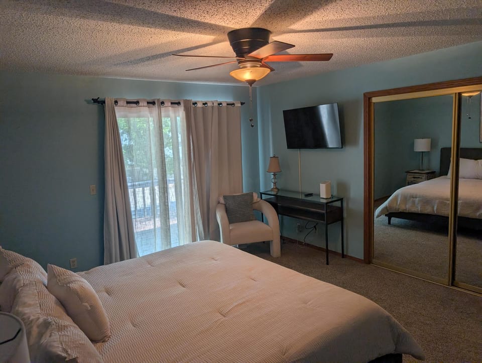 Sliding glass door with sheer curtains opens to a balcony, next to a white armchair and a wall-mounted TV.
