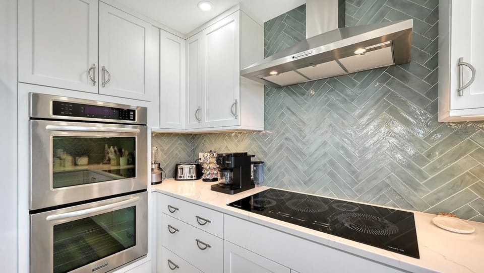 Stainless steel double oven beside white cabinetry and a glass-top induction cooktop with teal herringbone tile backsplash.