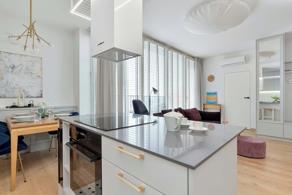 A contemporary kitchen island is equipped with an integrated stovetop and an oven built into the cabinetry below. The perspective looks past the cooking zone toward a bright dining area and large windows.