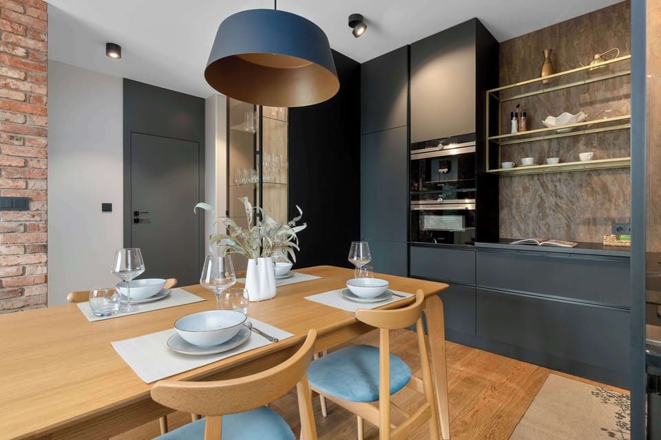 A wooden dining table is set with white plates, cloth napkins, and glassware under a blue pendant lamp. The matte black kitchen lines the background.