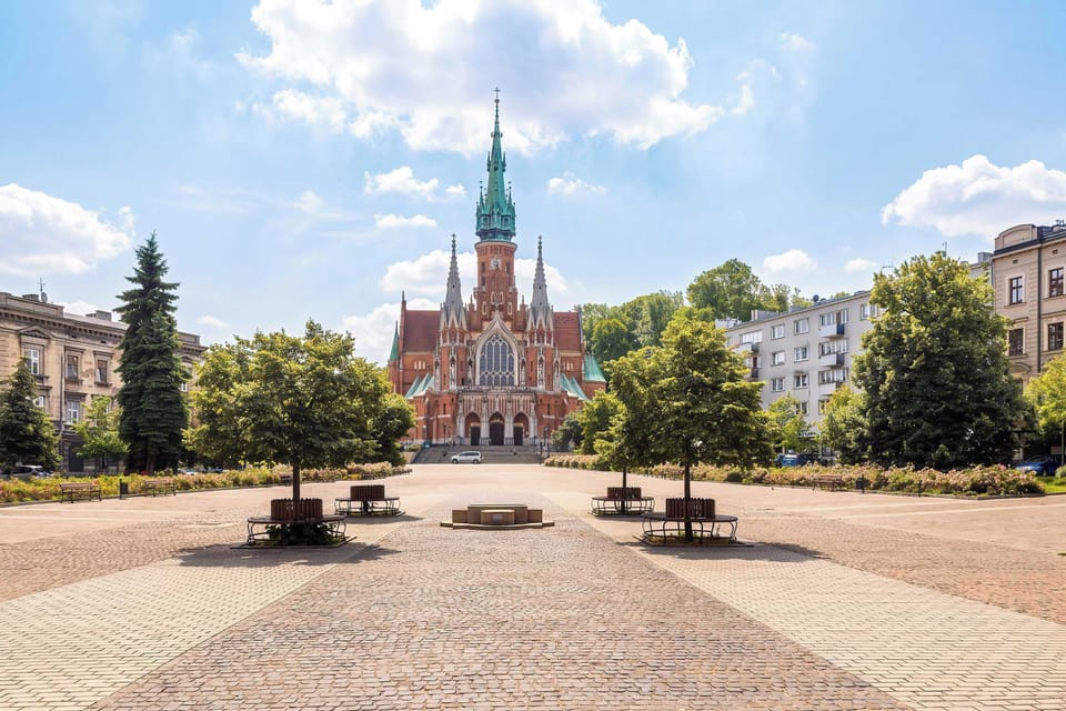 A wide brick path cuts through a green public square lined with young trees and benches. In the distance, a grand brick church with a tall spire stands prominently against a partly cloudy sky.