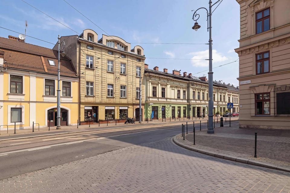 A cobblestone street curves gently alongside historic, multi-story buildings with classic architecture. A street lamp stands tall on the sidewalk under a bright blue sky.