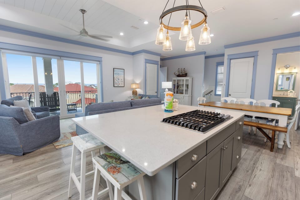 Your spacious kitchen and living area features a modern gas cooktop island, coastal-inspired decor, and bright natural light flowing through large windows overlooking the rooftops.