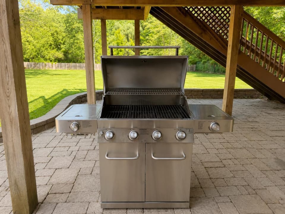 Stainless steel gas grill with five control knobs on a stone-paved patio under a wooden deck staircase.