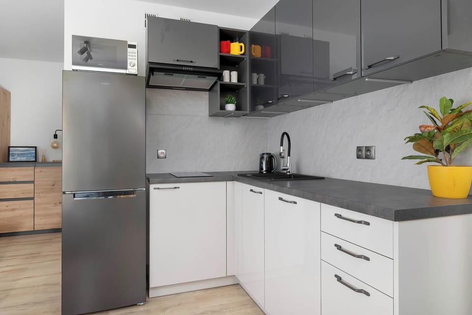 A contemporary kitchenette space showcases sleek dark grey lower cabinetry topped with light wooden countertops. A tall refrigerator stands at the end of the counter line under bright overhead lighting.