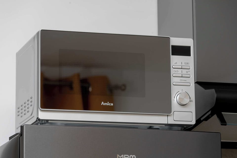 A close-up view focuses on a sleek silver microwave oven sitting neatly integrated onto a shelf. The reflective glass front shows a clear view of the surrounding room.