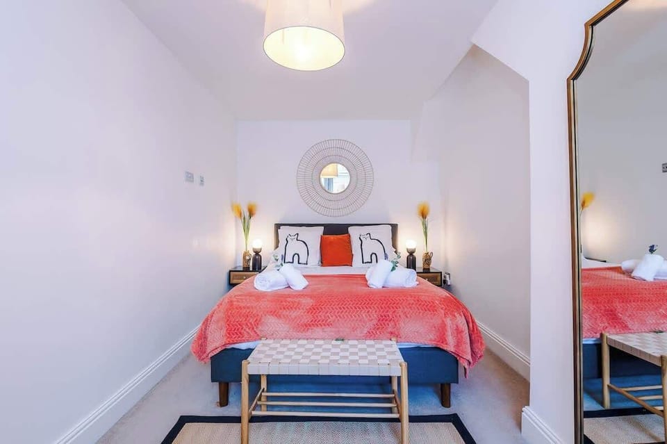 Cozy bedroom featuring a king-size bed with a coral bedspread, elephant-print cushions, and neatly folded swan towels. A round mirror above the headboard adds charm, while a gold-framed floor mirror and black clothing rack.