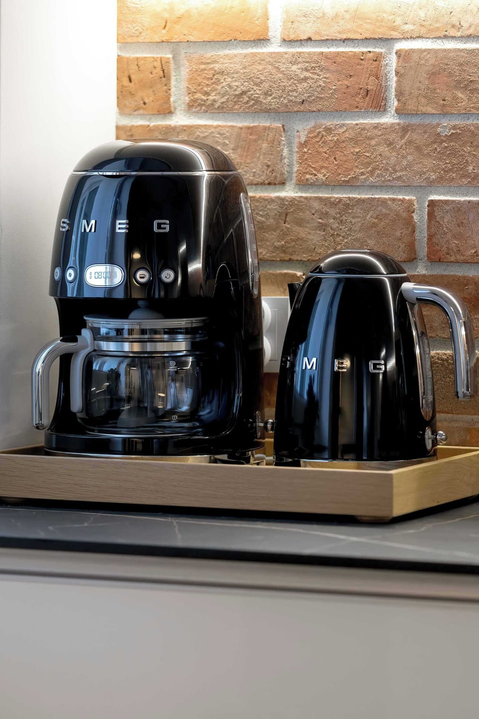 A close-up shot captures a sleek black air fryer and a matching electric kettle resting on a kitchen countertop. The background features a textured brick wall that enhances the contemporary aesthetic.
